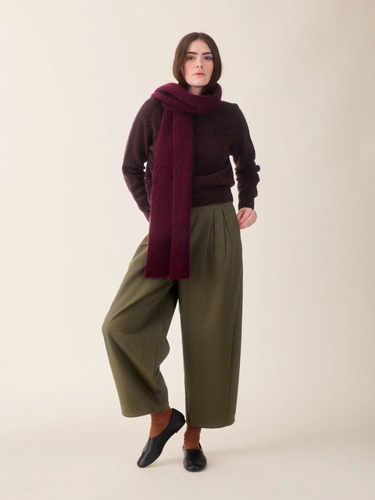A model stands against a plain beige studio background wearing a dark burgundy knit sweater, a matching dark burgundy knit scarf, olive green wide-leg cropped pants, and black slip-on shoes.