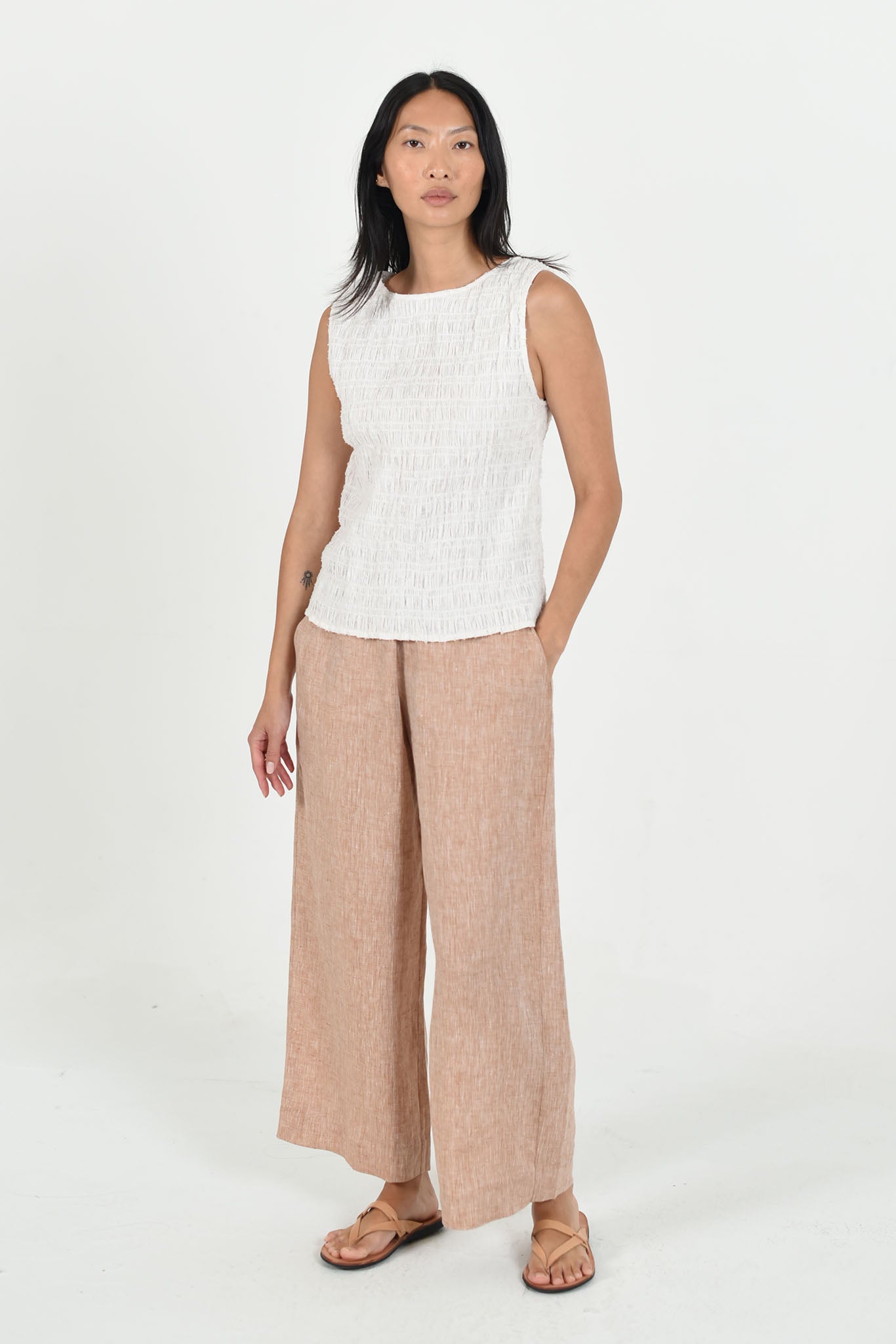 Female model with long black hair wearing a white vest top and wide leg biscuit colored linen pants