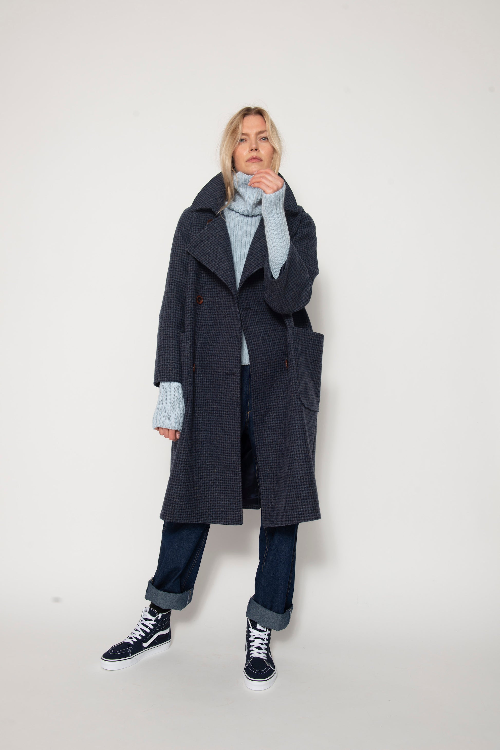 Woman standing wearing long blue houndstooth coat, pale blue sweater and baggy jeans