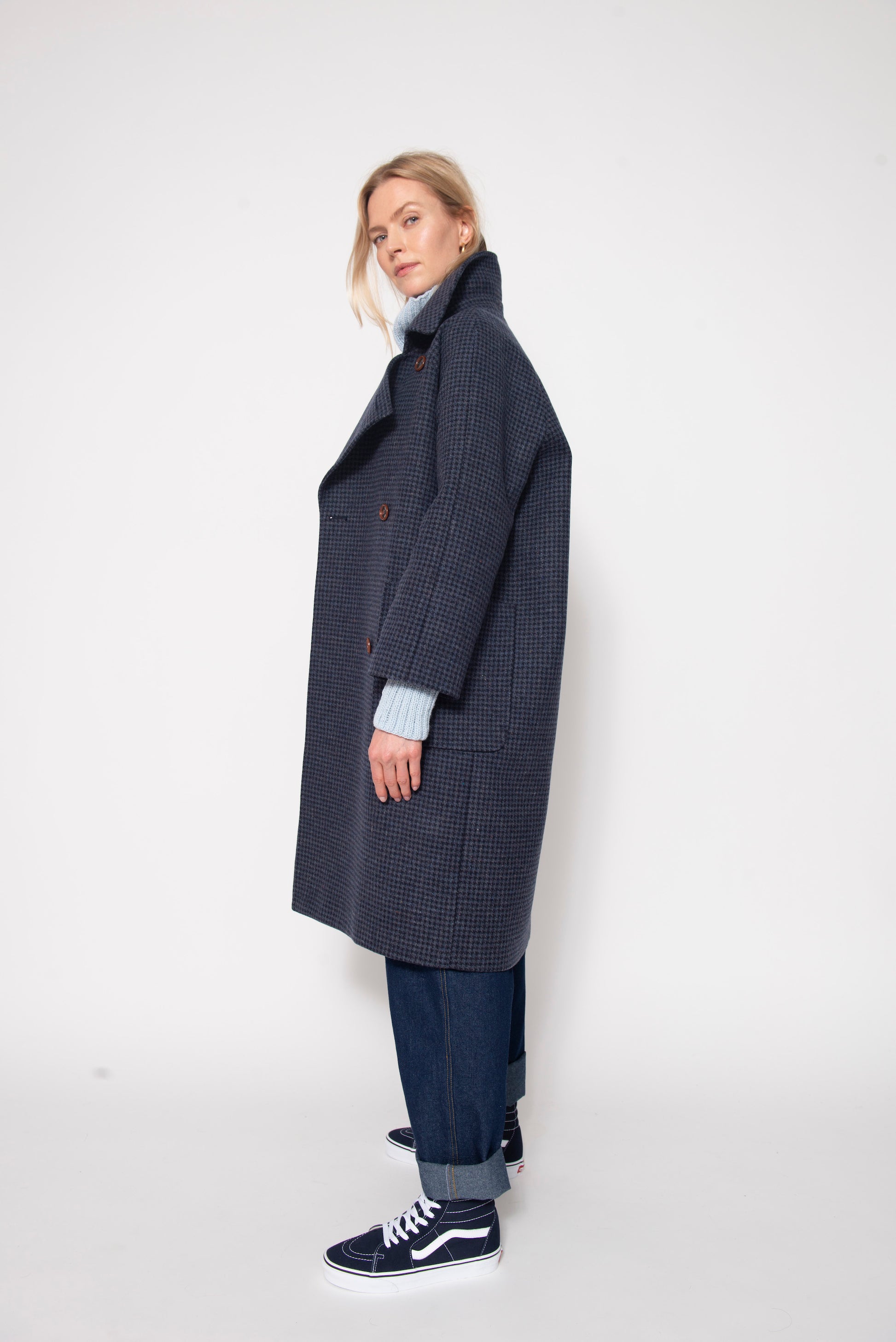 Woman standing wearing long blue houndstooth coat, pale blue sweater and baggy jeans