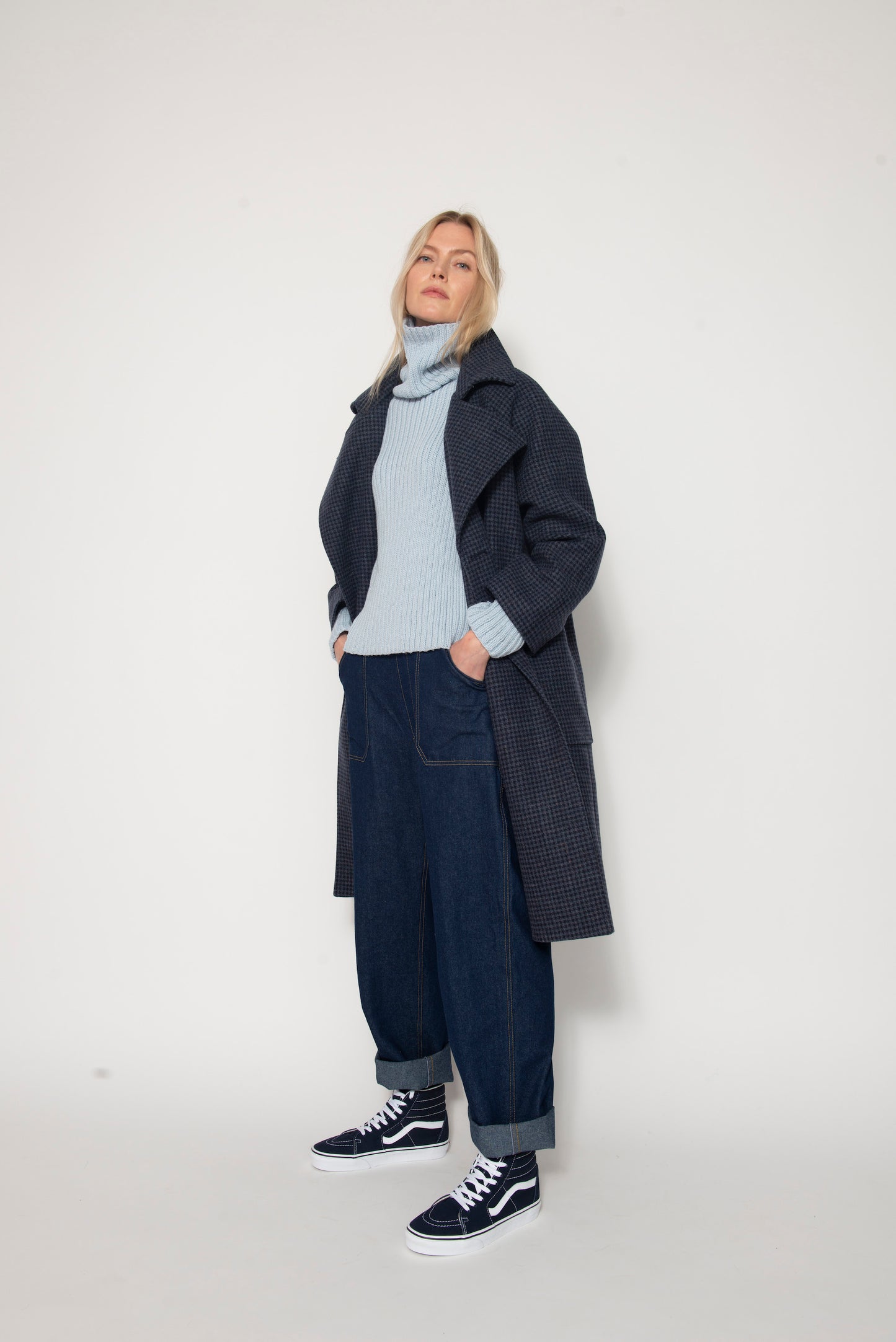 Woman standing wearing long blue houndstooth coat, pale blue sweater and baggy jeans